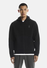 Musta vetoketjullinen huppari, valmistettu pehmeästä kankaasta, jossa etutasku, säädettävä huppu ja minimalistinen logo rinnassa. Rentoa istuvuutta.