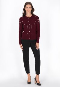 Bordowy kardigan z okrągłym dekoltem, ozdobnymi perełkami oraz zapięciem na guziki, zestawiony z czarnymi zwężającymi się spodniami i szpilkami.