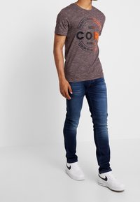 Grå melerat kortärmad T-shirt med cirkulär grafisk tryck i orange och svart. Tillsammans med blå denimjeans och vita sneakers.