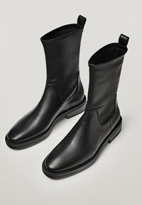 Bottines en cuir noir avec une texture lisse, un bout rectangulaire et des panneaux élastiques sur les côtés. Elles présentent un talon plat et des languettes d'enfilage.
