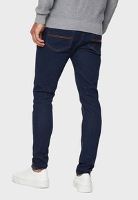 Hombre vestido con jeans ajustados de color azul oscuro y sneakers blancos, de pie con la espalda hacia el espectador contra un fondo liso.