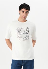T-shirt en coton blanc avec un design graphique du Mont Fuji entouré de fleurs et de texte asiatique. Col ras du cou et manches courtes.