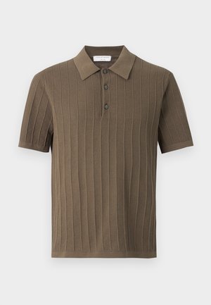 Fiatal férfi göndör hajjal, barna rövid ujjú kötött pólót, széles szárú kék farmert és fehér tornacipőt viselve áll, egyszerű háttér előtt.