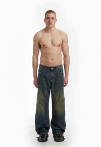 Posete blå denimjeans med et distinkt splittedesign, falmede seksjoner og kontrasterende gult søm, kombinert med svarte sneakers.