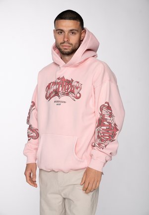 Mann mit kurzen Haaren, der einen pinken Hoodie mit rotem grafischem Text auf Brust und Ärmeln sowie beigefarbene Hosen trägt, steht vor weißem Hintergrund.