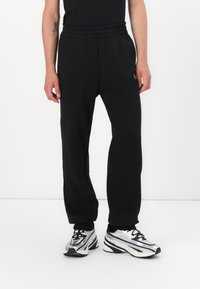Sorte sweatpants med elastisk talje, sidelommer, tapered ben og en glat overflade. Bæres med sølvfarvede træningssko.