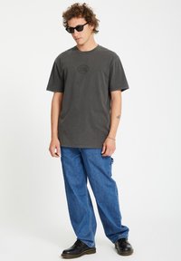 T-shirt di cotone grigio scuro con collo rotondo e maniche corte, caratterizzato da un logo centrale. Abbinato a pantaloni in denim largo blu e scarpe nere.