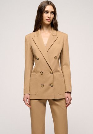 Tan double-breasted blazer with large button closures, notch lapels, and two front pockets. Tailored fit and smooth texture.