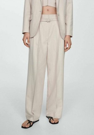 Persoon draagt beige high-waist wijde broek met bijpassende cropped blazer en zwarte teenslippers tegen een effen achtergrond.