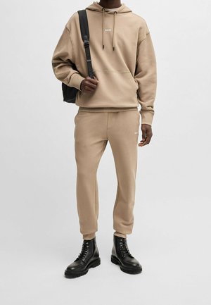 Man draagt een beige BOSS-hoodie en bijpassende joggingbroek, zwarte veterlaarzen en draagt een zwarte schoudertas.