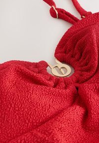 Top da bagno in tessuto rosso strutturato con dettaglio di chiusura circolare dorata e spalline regolabili, disteso su una superficie bianca.