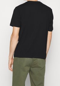 Camiseta negra de mangas cortas con cuello redondo, hecha de algodón suave. La parte trasera es lisa y tiene un corte holgado, combinada con pantalones verde oliva.