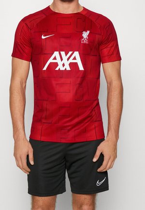 Mężczyzna w czerwonej koszulce piłkarskiej Liverpool FC z białymi logo AXA i Nike, w czarnych szortach Nike, stojący na jednolitym tle.