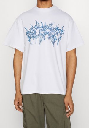 Personne portant un T-shirt blanc oversize avec un motif graphique abstrait bleu et un pantalon cargo vert olive, se tenant devant un fond uni.