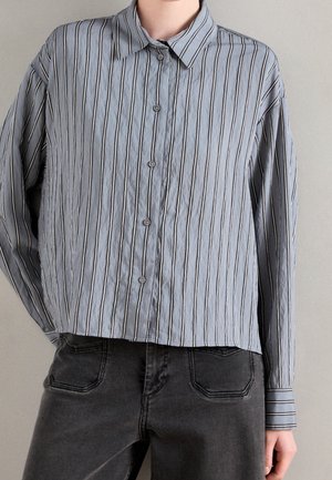 Person trägt ein hellblaues, langärmliges Hemd mit vertikalen weißen und schwarzen Streifen sowie schwarze Jeans.