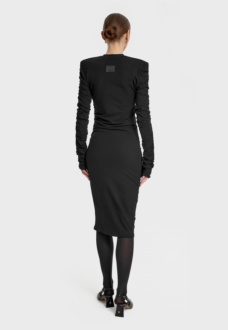 Czarna sukienka z długim rękawem o dopasowanej sylwetce, z teksturowanej tkaniny i detalami na ramionach. Ma długość poniżej kolan i wysokie obcasy.