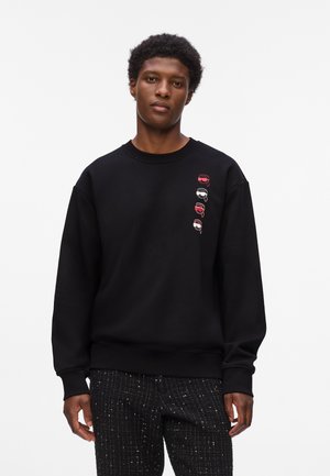 Zwarte sweatshirt met een ronde hals, voorzien van kleine gekleurde afbeeldingen van gezichten met verschillende hoofdbanden aan de linkerkant. Zacht stof, ontspannen pasvorm.