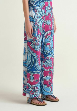 Weit geschnittene Hosen mit lebendigen pinken, blauen und weißen Paisley-Mustern, kombiniert mit offenen Sandalen mit Perlenbesatz an nackten Füßen.