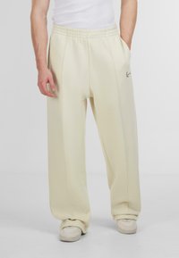 Pantaloni de trening de culoare crem deschis, cu o croială relaxată, talie elastică, buzunare laterale și un mic detaliu cu logo negru pe coapsă.