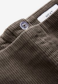 Brown corduroy fabric with vertical ribbed texture, featuring a clear button closure and label reading "NEXT." Close-up view of waistband.