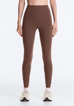 Vrouw draagt bruine leggings met hoge taille en witte sneakers, staand rechtop tegen een effen witte achtergrond.