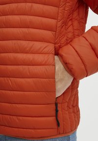 Polstret orange jakke med quiltning, elastiske cuffs og en lynlåslomme med en snor til justering. Teksturen virker glat.