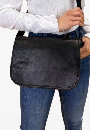 Black leather shoulder bag with a smooth texture, rounded edges, and an adjustable strap, held by a person wearing a white top and jeans.