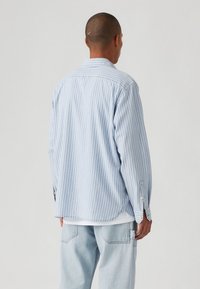 Camicia a maniche lunghe a righe azzurro chiaro e bianco, con orlo curvo e dettagli con polsini abbottonati, realizzata in tessuto morbido, indossata sopra pantaloni di jeans chiari.