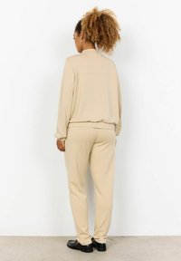 Beige loungewear set with long-sleeve top and pants, featuring a relaxed fit and ribbed cuffs and waistband. Black footwear completes the look.