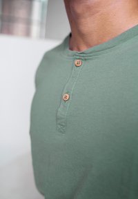 Zelená henley košile vyrobená z měkké látky, která má zaoblený výstřih, tři dřevěné knoflíky a hladkou strukturu.