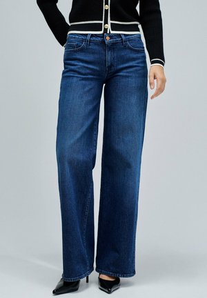 Mujer que lleva puestos unos pantalones de pierna ancha azul oscuro, tacones negros de punta, y un cárdigan negro con ribete blanco y botones dorados.