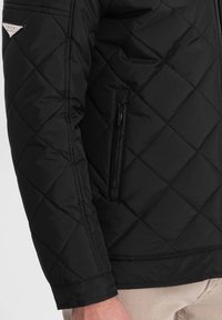 Sort quiltet jakke med en blank finish, lynlås lomme på siden og en sølvfarvet logoaccent på den øverste ærme.