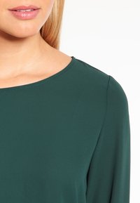 Top verde in tessuto liscio con scollatura rotonda. Caratterizzato da un design pulito e minimale senza decorazioni. Tessuto morbido e maniche aderenti.
