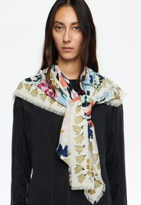 Bufanda de tela suave con estampado floral, que presenta flores azules, rosas y amarillas sobre un fondo claro; con bordes con flecos; colocada alrededor del cuello.
