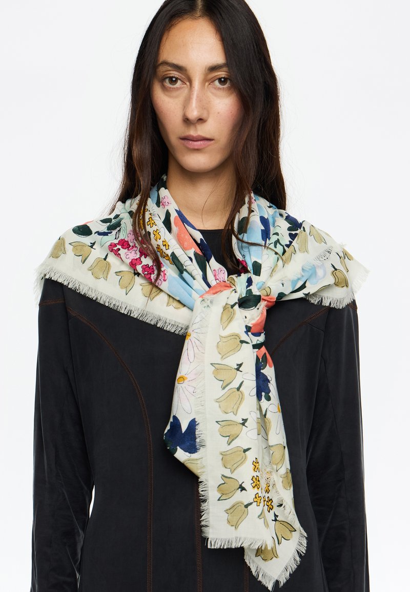 Bufanda de tela suave con estampado floral, que presenta flores azules, rosas y amarillas sobre un fondo claro; con bordes con flecos; colocada alrededor del cuello.