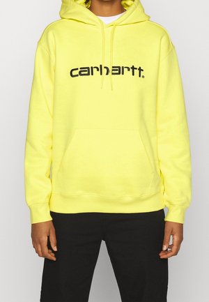 Personne portant un sweat à capuche Carhartt jaune vif avec un texte noir et un pantalon noir, debout devant un fond uni.