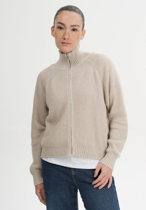 Woman with pulled-back hair wearing a beige ribbed zip-up sweater, white shirt underneath, and blue jeans against a plain background.