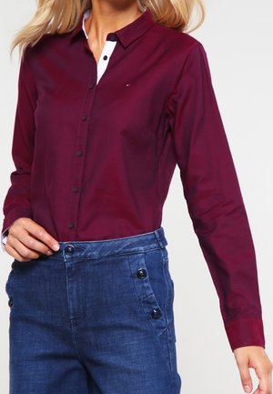 Femme portant une chemise bordeaux à manches longues boutonnée, rentrée dans un pantalon bleu taille haute avec des boutons latéraux.