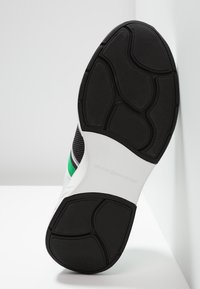 Czarnobiała podeszwa sportowego buta z teksturowanym gumowym bieżnikiem, z zielon�ą i srebrną linią akcentującą po boku oraz zakrzywionymi elementami wzornictwa.