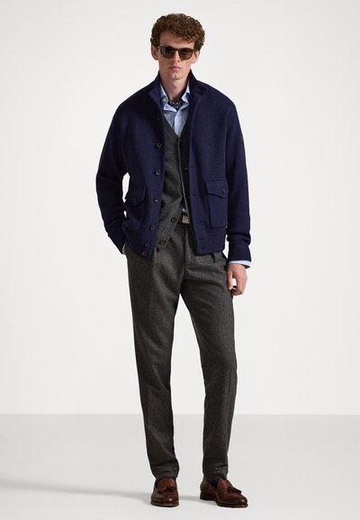 Mornarsko pleteno jopico z visokim ovratnikom, gumbi in dvema žepoma, oblečeno čez sivo telovnik in svetlobluzo srajco, v kombinaciji s sivimi hlačami.