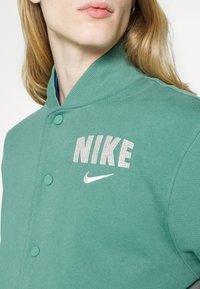 Grön sweatshirtjacka med ribbat krage, prydd med vit "NIKE" broderi och en liten Nike swoosh. Två knappknäppningar synliga.