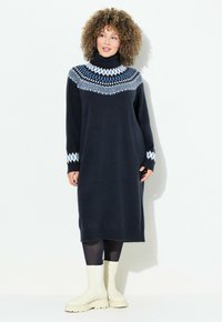 Abito lavorato a maglia blu navy con collo alto, con un design a motivo blu e bianco sulla scollatura e sui polsini. Indossato con stivali bianchi alla caviglia.