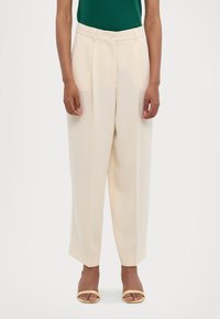 Krémové široké kalhoty s předními sklady, vysokým pasem a bočními kapsami, doplněné zeleným topem a žlutými sandály na podpatku.