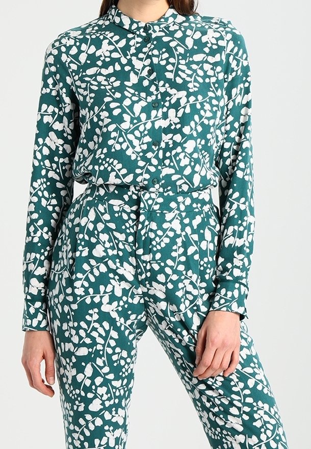 Chemise boutonnée turquoise et pantalon assorti avec un motif de feuilles et branches blanches, porté par une personne se tenant devant un fond clair uni.