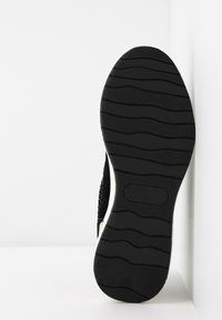 Semelle de chaussure noire avec motifs ondulés, affichée verticalement sur fond blanc, montrant les détails du design de l'adhérence.