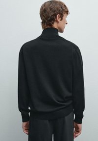 Pullover nero lavorato a maglia con colletto alto e polsini a costine. Presenta una texture liscia e una vestibilità comoda, visto dari retro.
