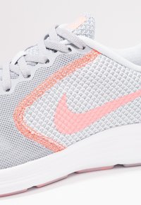 Lichtgrijze mesh sneaker met een roze Nike swoosh, oranje stiksels en een witte zool, met gestructureerde oppervlakte details.