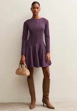 Vestido ajustado de manga larga en púrpura con falda acampanada, combinado con botas altas beige hasta la rodilla y un pequeño bolso beige. Tela suave con detalles de costura.