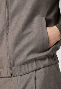 Grau strukturierte Jacke mit einem Reißverschluss vorne und seitlicher Tasche. Verfügt über einen gerafften Taillenbund und gerippte Bündchen. Das Material wirkt leicht.