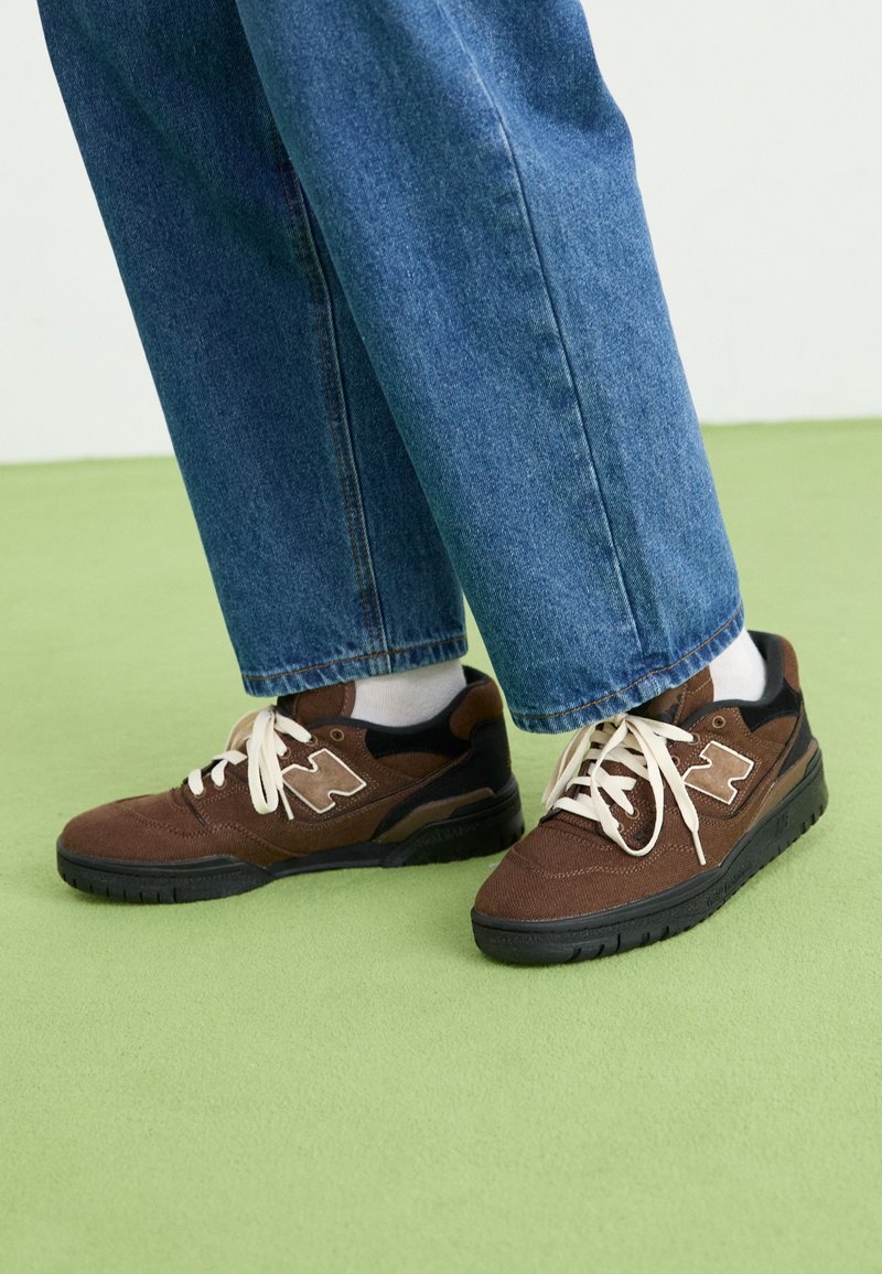 Zapatillas de ante marrón con acentos negros y un logo blanco, que cuentan con cordones blancos y una suela de goma negra texturizada. Combinadas con pantalones de mezclilla azules.